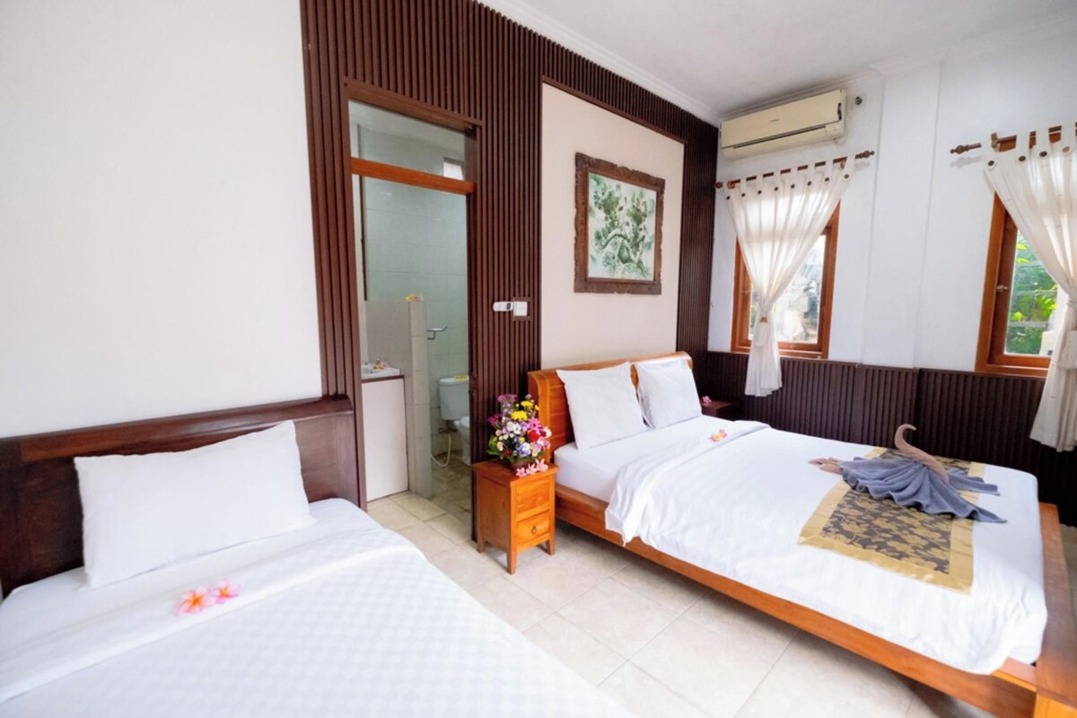 A hotel room with two beds, a wooden headboard, a bedside table with a vase of flowers, a window with curtains, and an air conditioning unit.