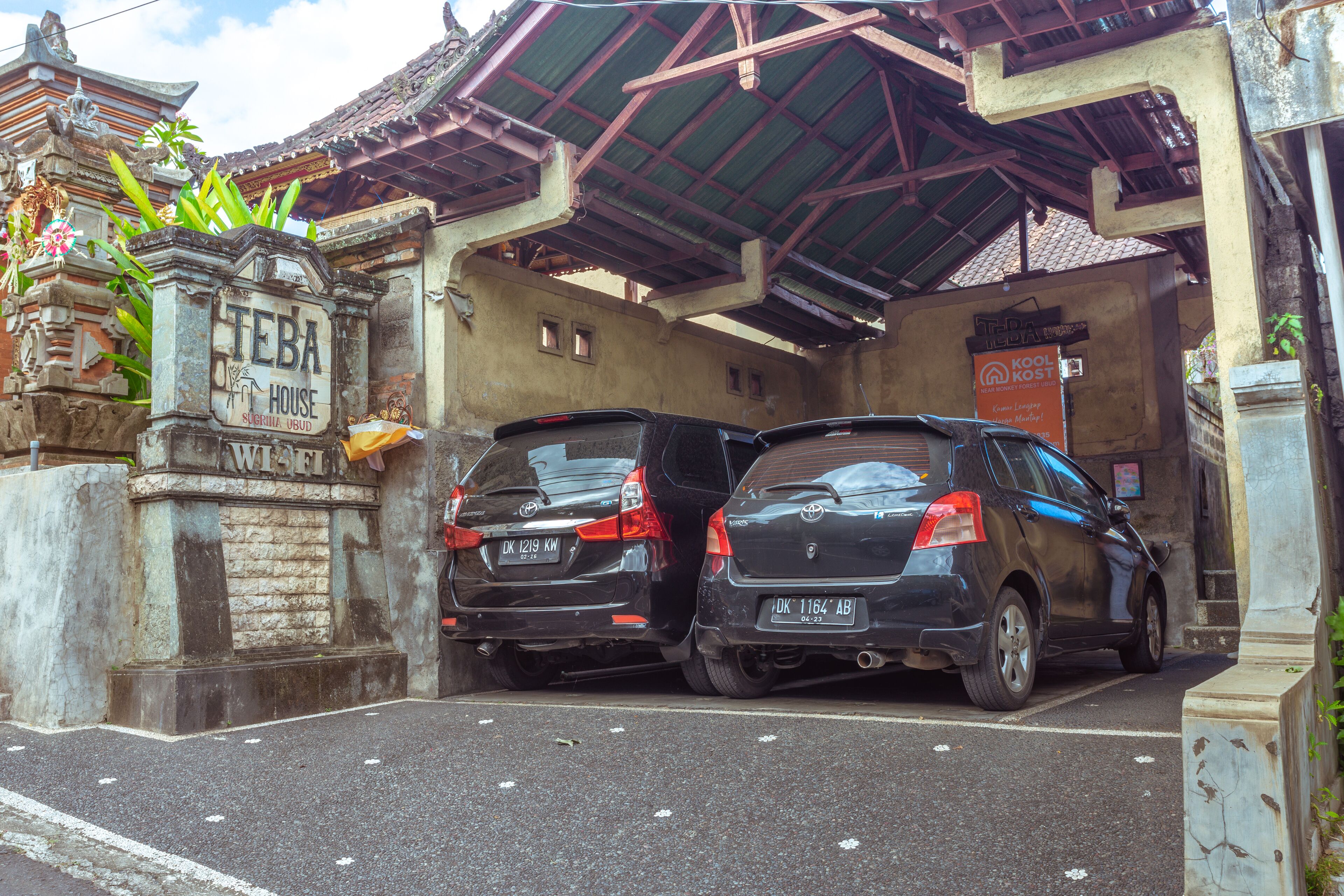 Un espace de stationnement où deux voitures sont garées devant un bâtiment doté d’un toit traditionnel et d’une enseigne indiquant « TEBA HOUSE ».