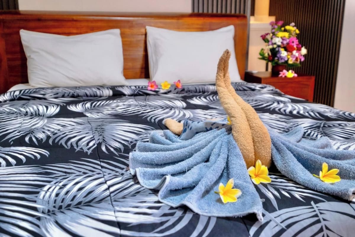 Una cama con una colcha estampada, una toalla doblada decorativamente y un florero con flores sobre una mesita de noche.