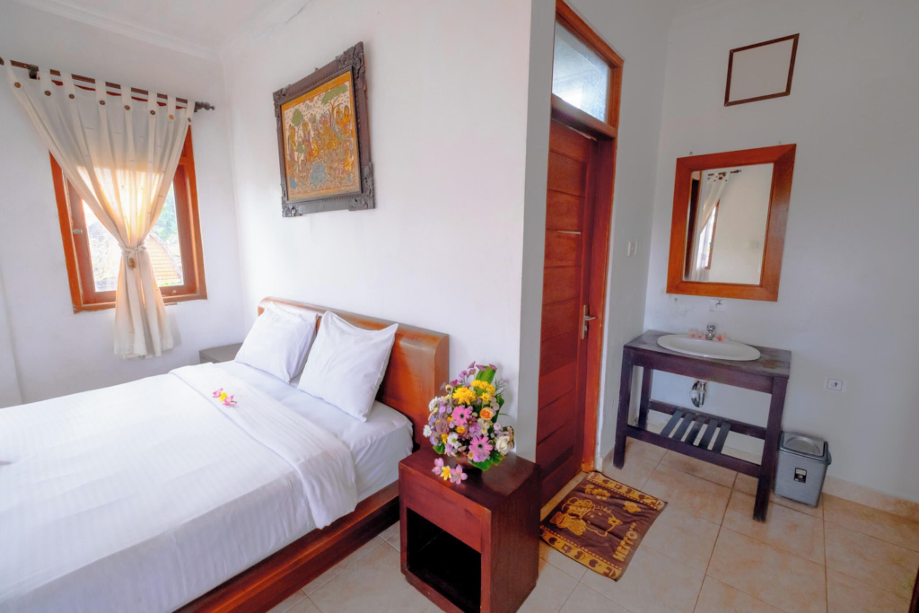 A bedroom with a bed, a nightstand with a vase of flowers, a wooden door, a bathroom with a sink and mirror, and a tiled floor.