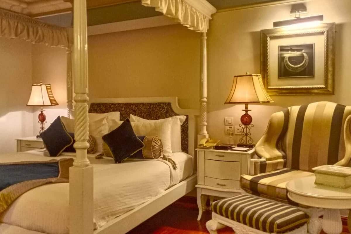 A four-poster bed with a canopy, a bedside table with a lamp, a striped armchair, and a small round table with a book.