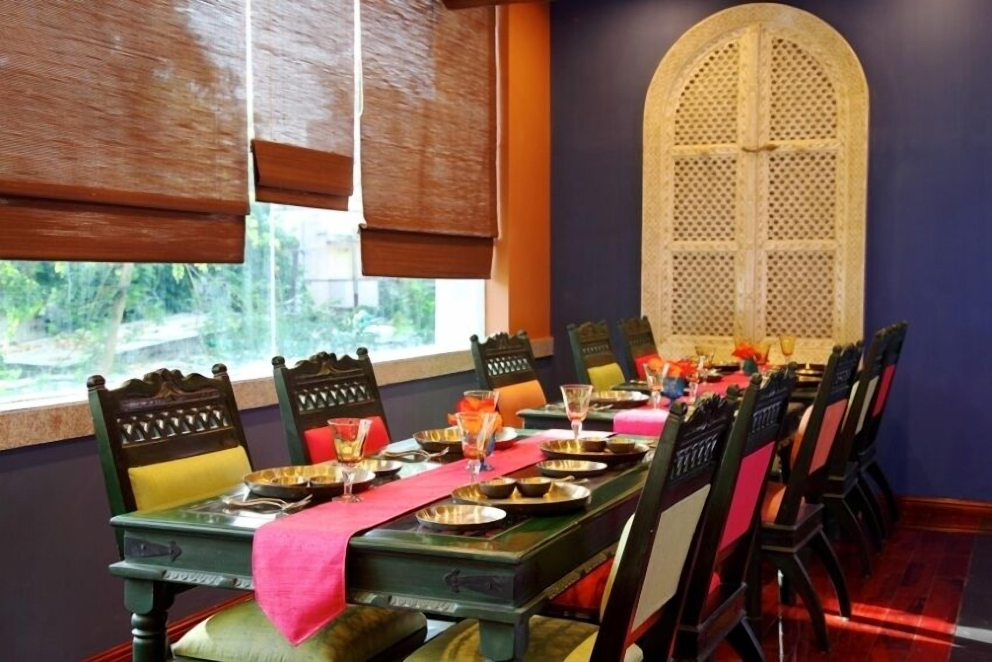 A dining area with a long table set for a meal, chairs with cushions, a decorative window with a lattice design, and a view of greenery outside.