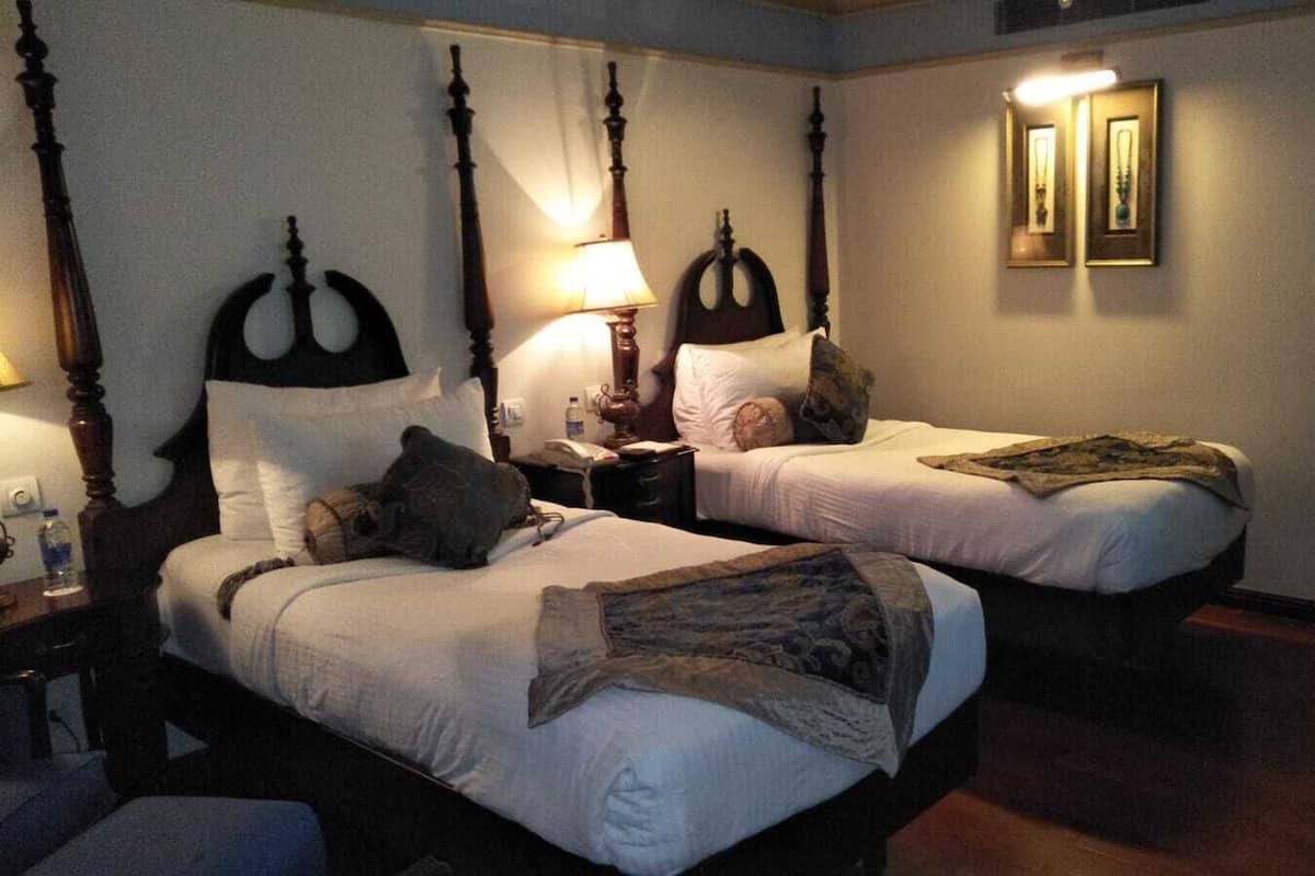 A hotel room with two beds, each with a wooden headboard and white bedding. There is a bedside table with a lamp and a bottle of water. The room has a wooden floor and a door leading to another area.
