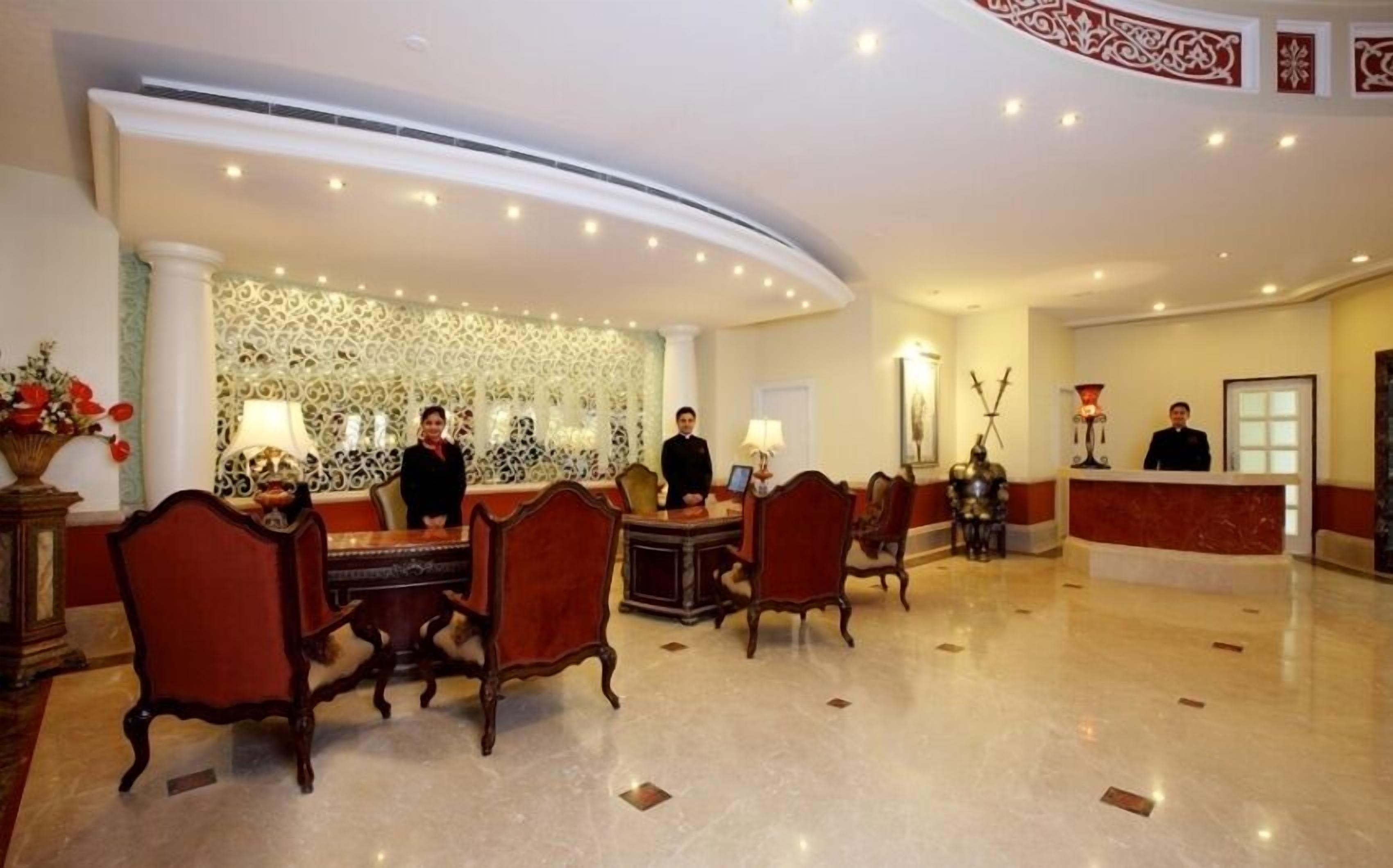 Um saguão de hotel com balcão de recepção, várias cadeiras, uma parede decorativa e piso de mármore.