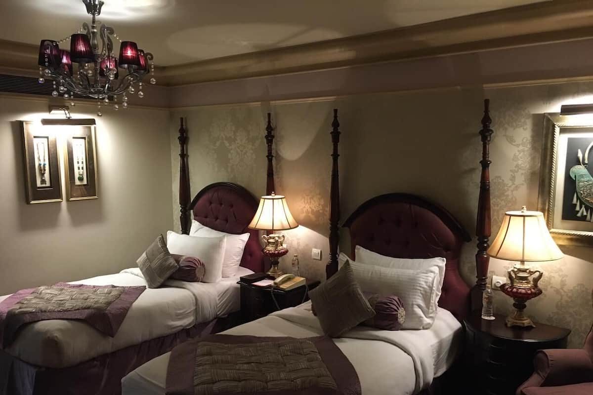 A hotel room with two beds, each with a headboard, bedside tables with lamps, and a chandelier.