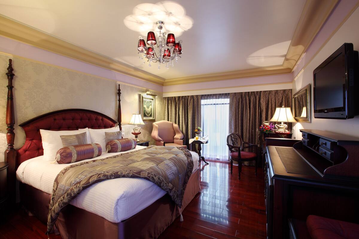 A hotel room with a large bed, a desk, a chair, a television, and a chandelier.