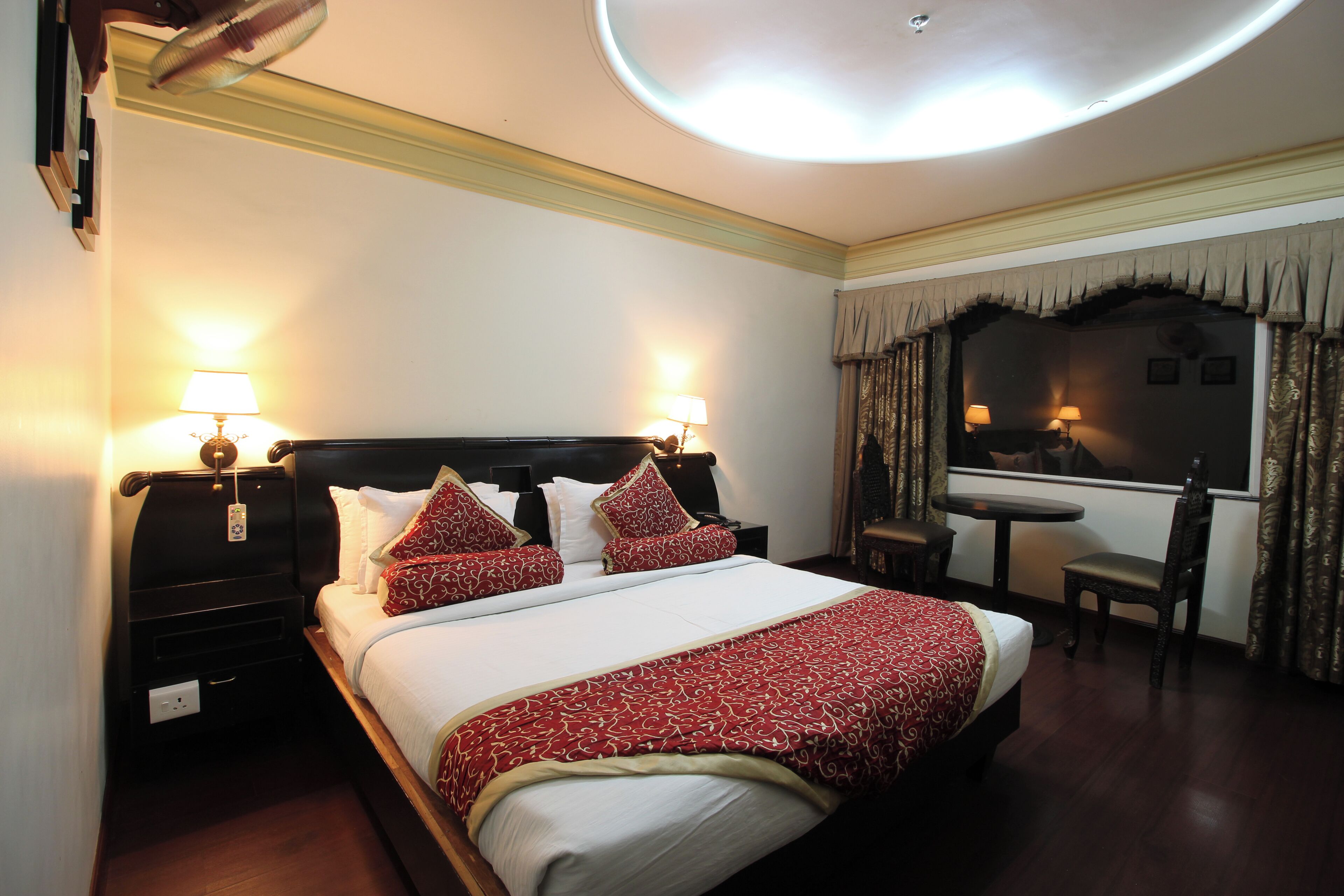 Habitación de hotel con una cama grande, dos mesitas de noche con lámparas, una pequeña mesa con una silla y un espejo sobre la cama.