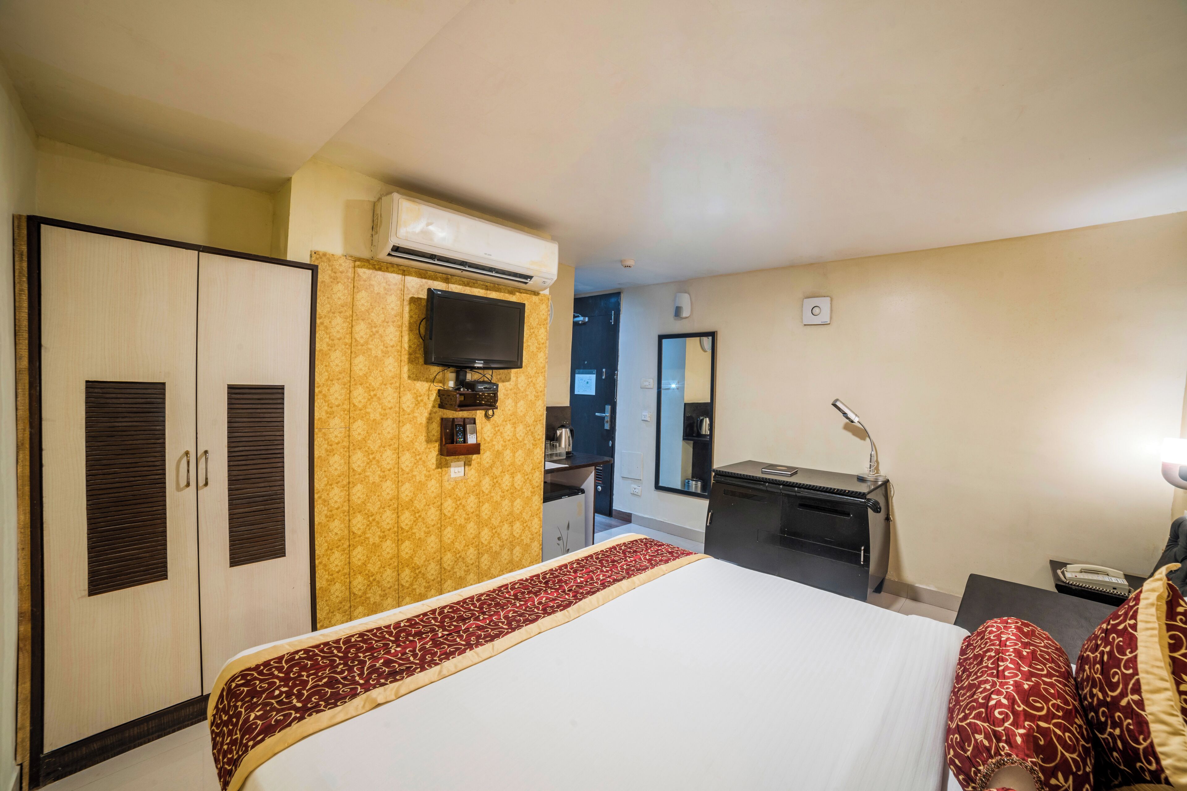 A hotel room with a bed, wardrobe, desk, mirror, and an air conditioning unit.
