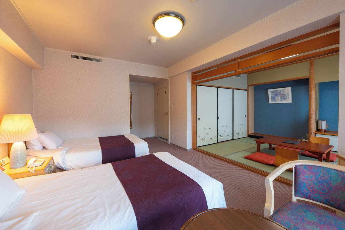 A hotel room with two beds, a desk, a chair, a lamp, and a sliding door leading to a traditional Japanese room.