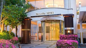 L’extérieur de l’hôtel Merveille ARIMA, avec son entrée en verre, est entouré de verdure et de fleurs roses.
