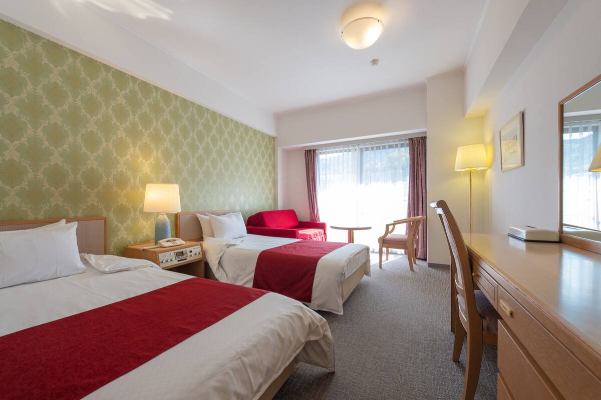 A hotel room with two beds, a desk, a chair, a lamp, and a window with curtains.