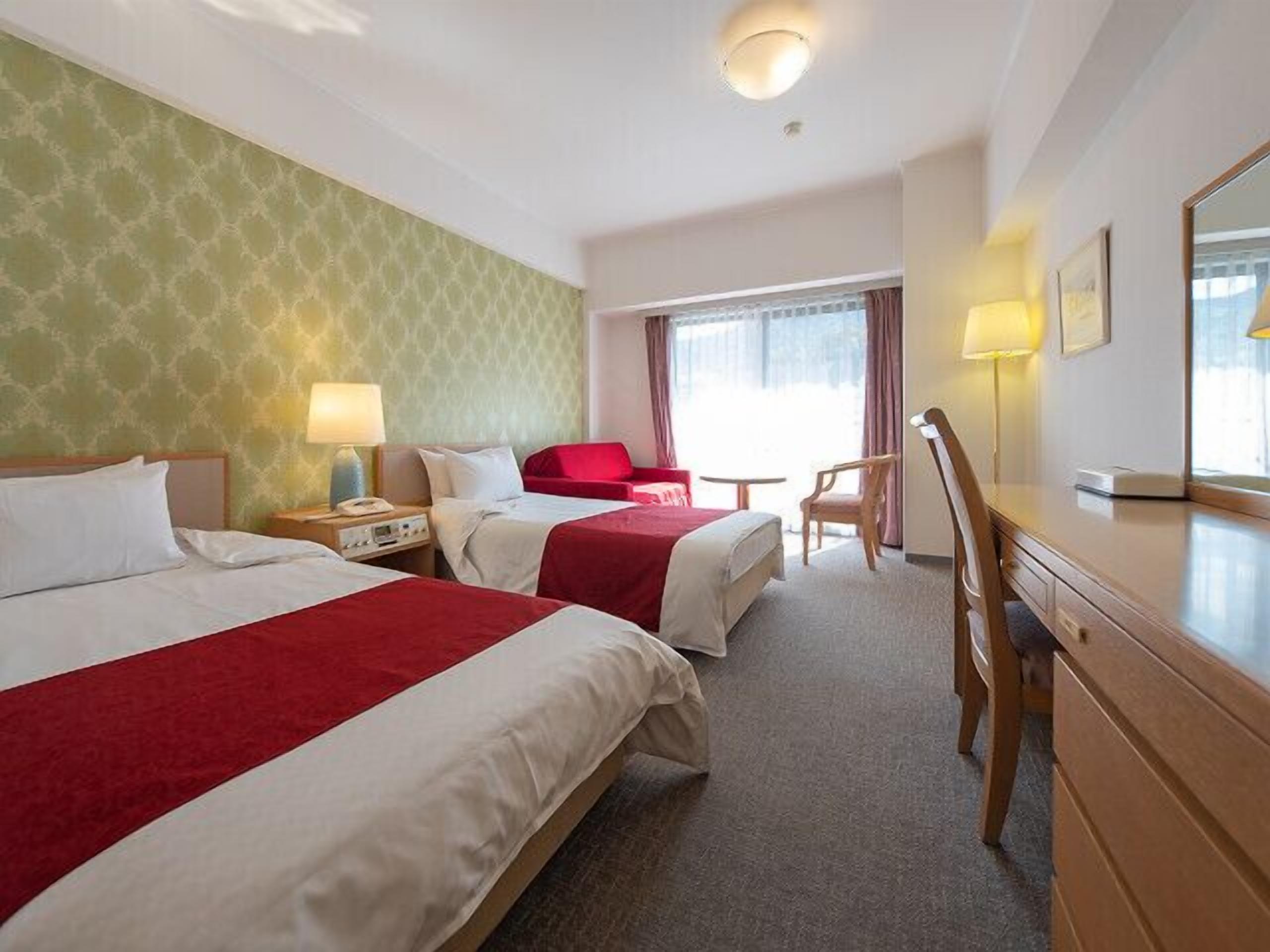 A hotel room with two beds, a desk, a chair, a lamp, and a window with curtains.