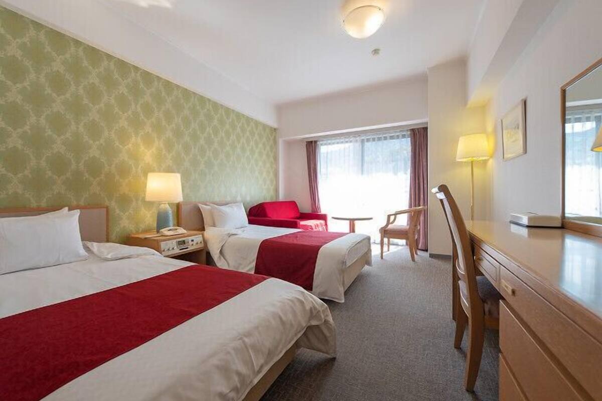 A hotel room with two beds, a desk, a chair, a lamp, and a window with curtains.