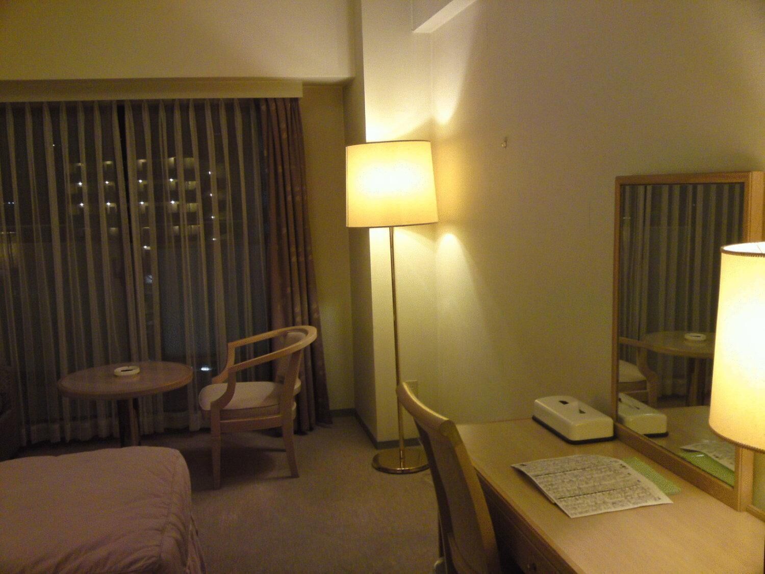 A hotel room with a bed, a desk with a lamp, a chair, a small table, and a window with curtains.