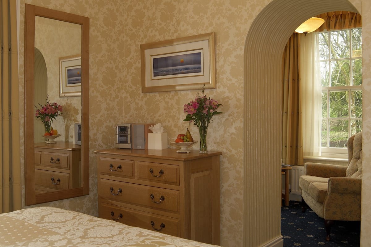 Quarto de hotel com um espelho grande, um quadro emoldurado, penteadeira de madeira com gavetas, mesa de cabeceira com um vaso de flores e uma janela com cortinas.