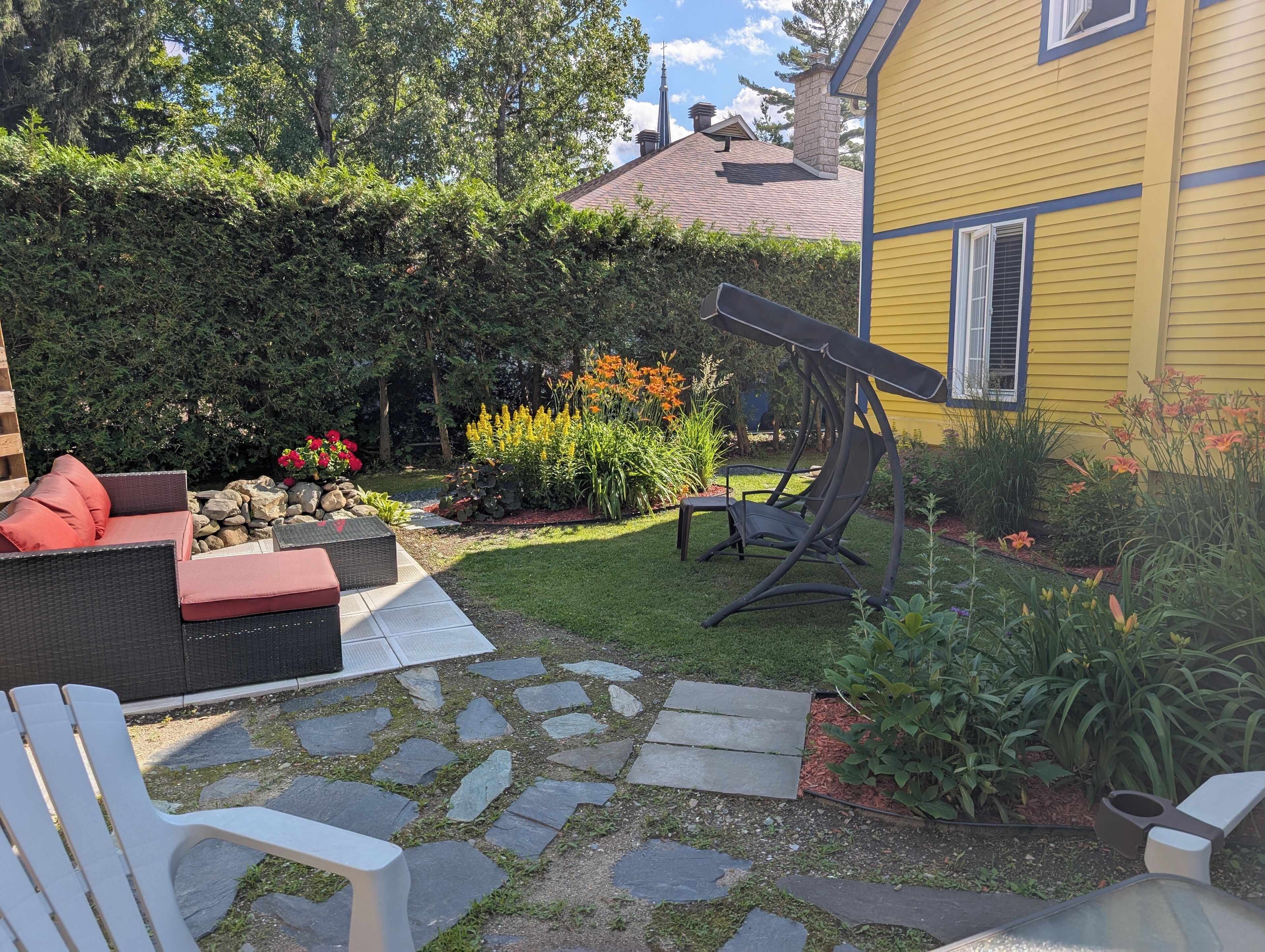 Um pátio com mobiliário de exterior, uma cadeira de balanço e um jardim com diversas plantas e flores.