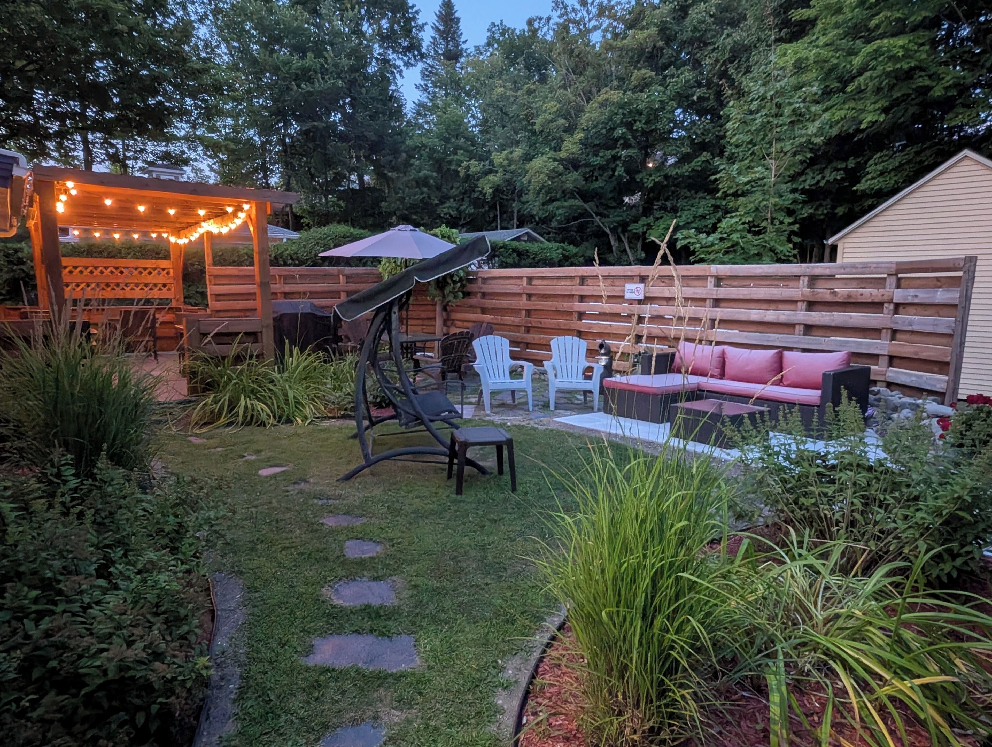 Een achtertuin met een houten terras, buitenmeubels en een pergola met lichtslingers.