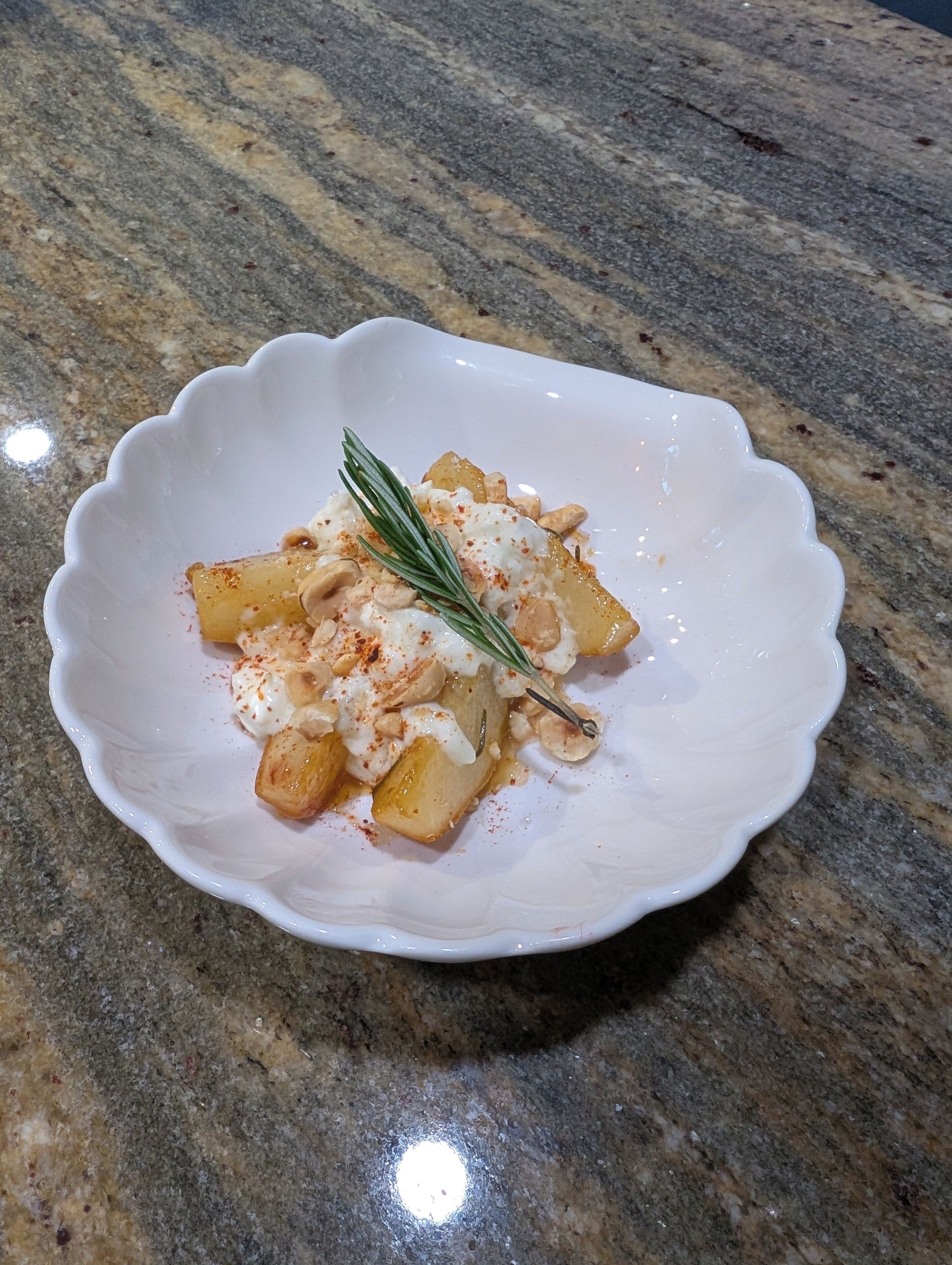 A white bowl with a scalloped edge containing a dish of food, possibly potatoes, with a sprig of rosemary and a sprinkle of red seasoning on a granite countertop.