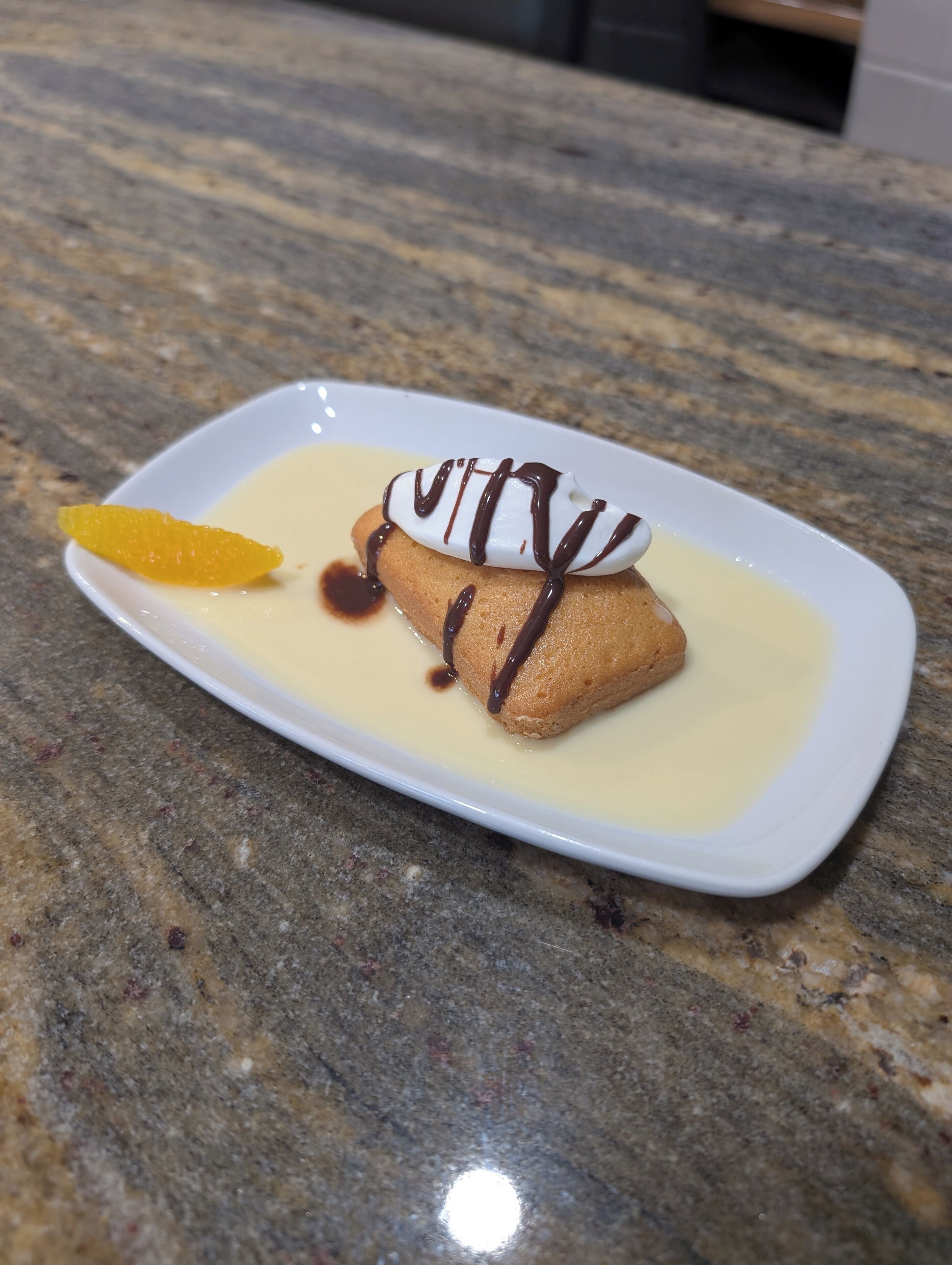 Dessert op een wit bord met chocoladesaus en een sinaasappelschijfje, op een granieten aanrecht.