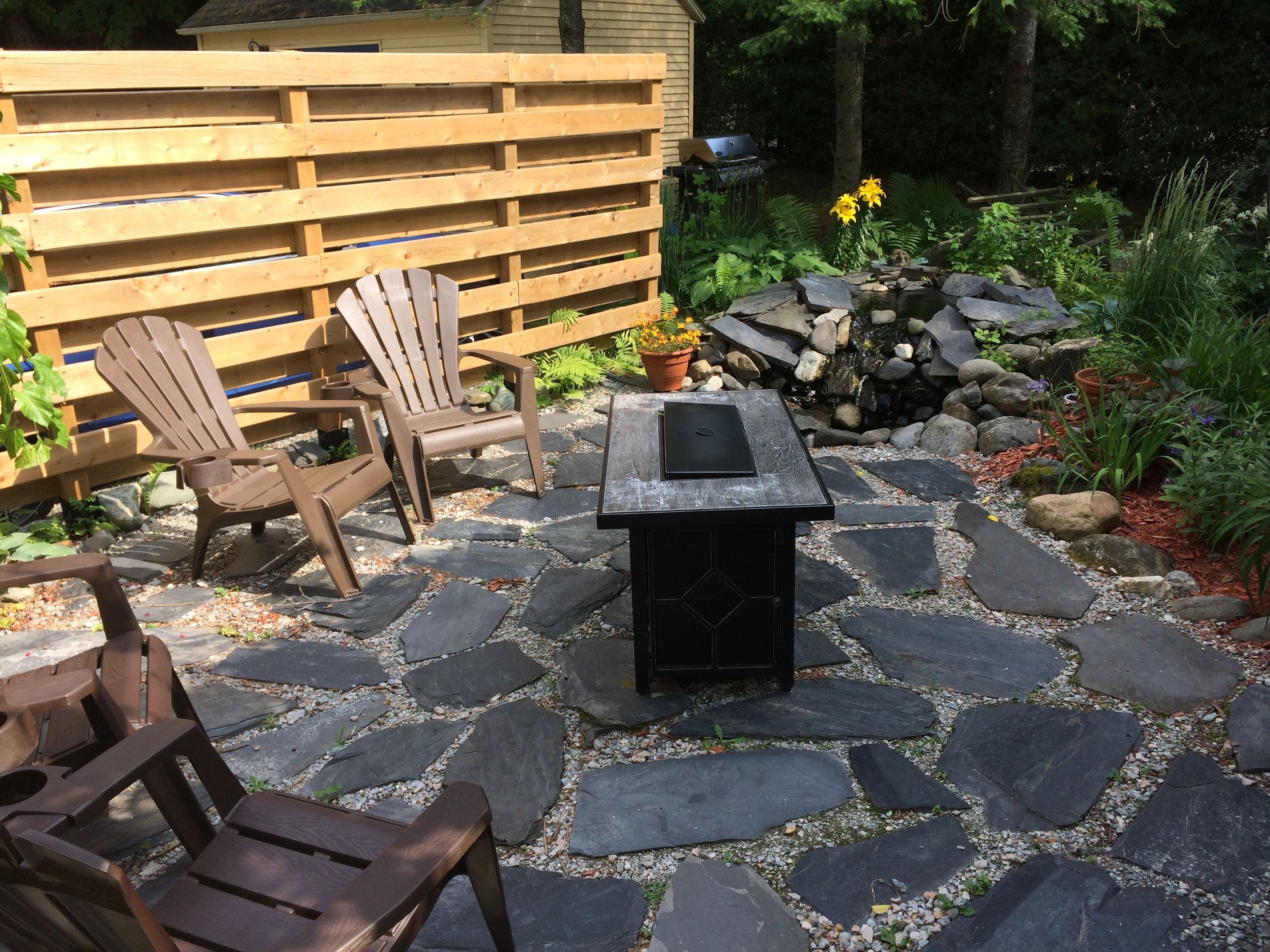 A patio with stone flooring, wooden chairs, a table, and a grill.
