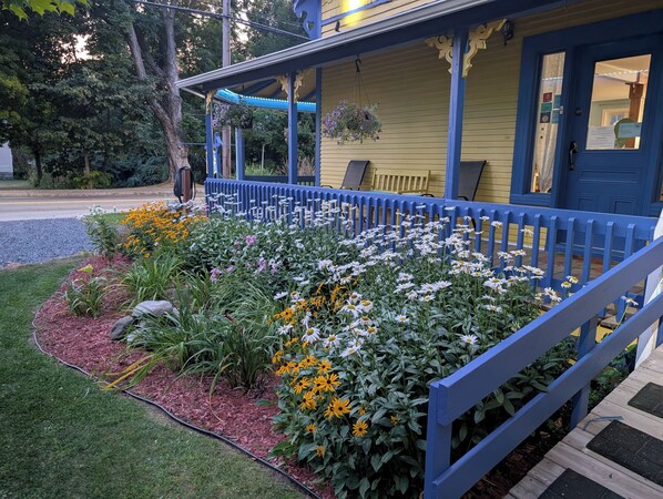 A porch with a blue railing, a yellow building, and a garden with various flowers. - Au Saut du Lit (Magog)
