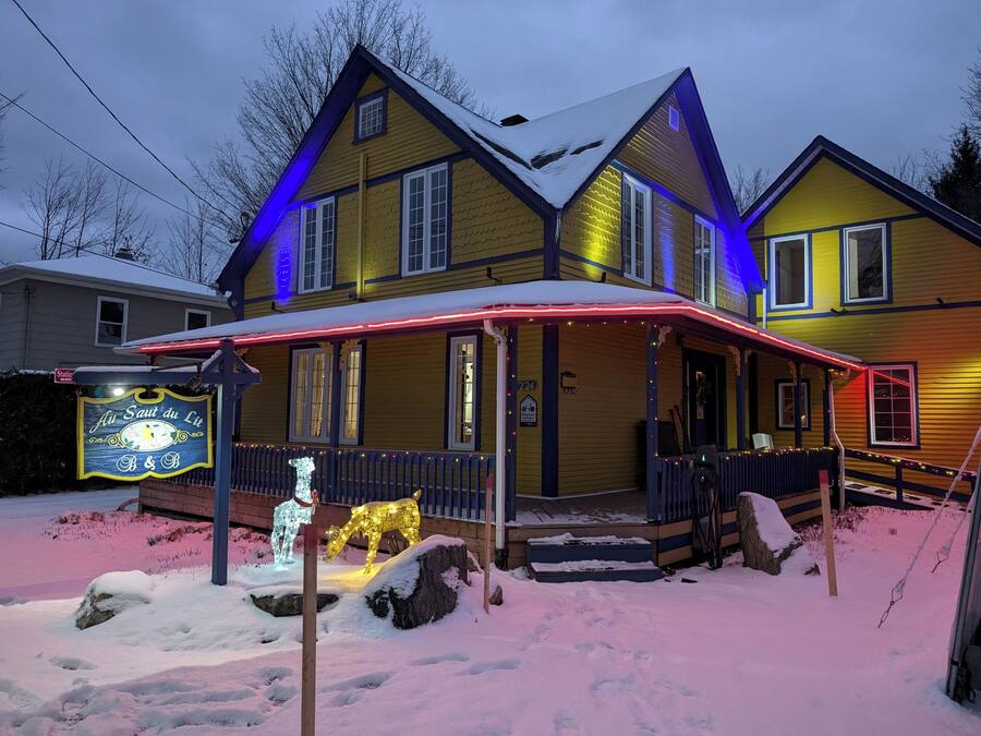 A two-story yellow house with a porch, snow on the ground, and illuminated decorations.