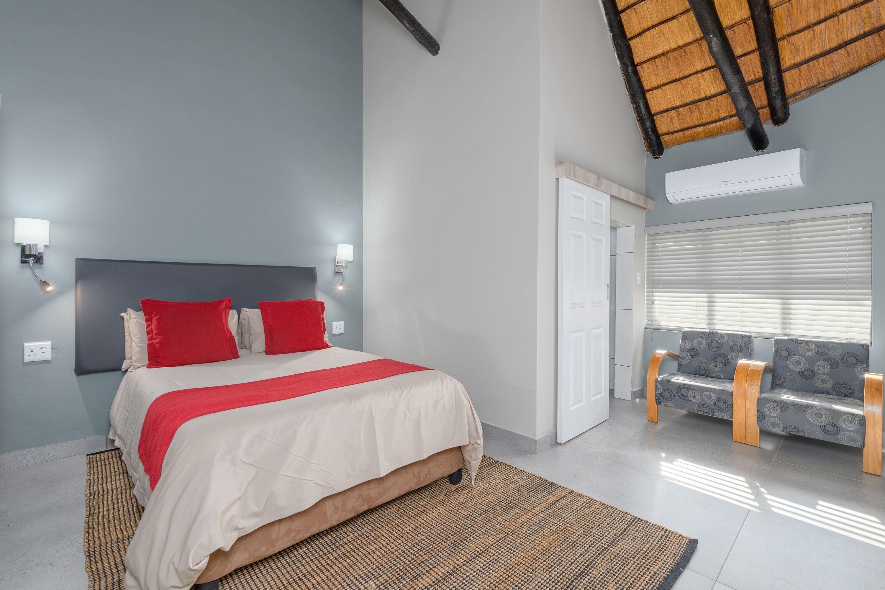 A bedroom with a bed, a sofa, a wall-mounted air conditioner, and a window with blinds.