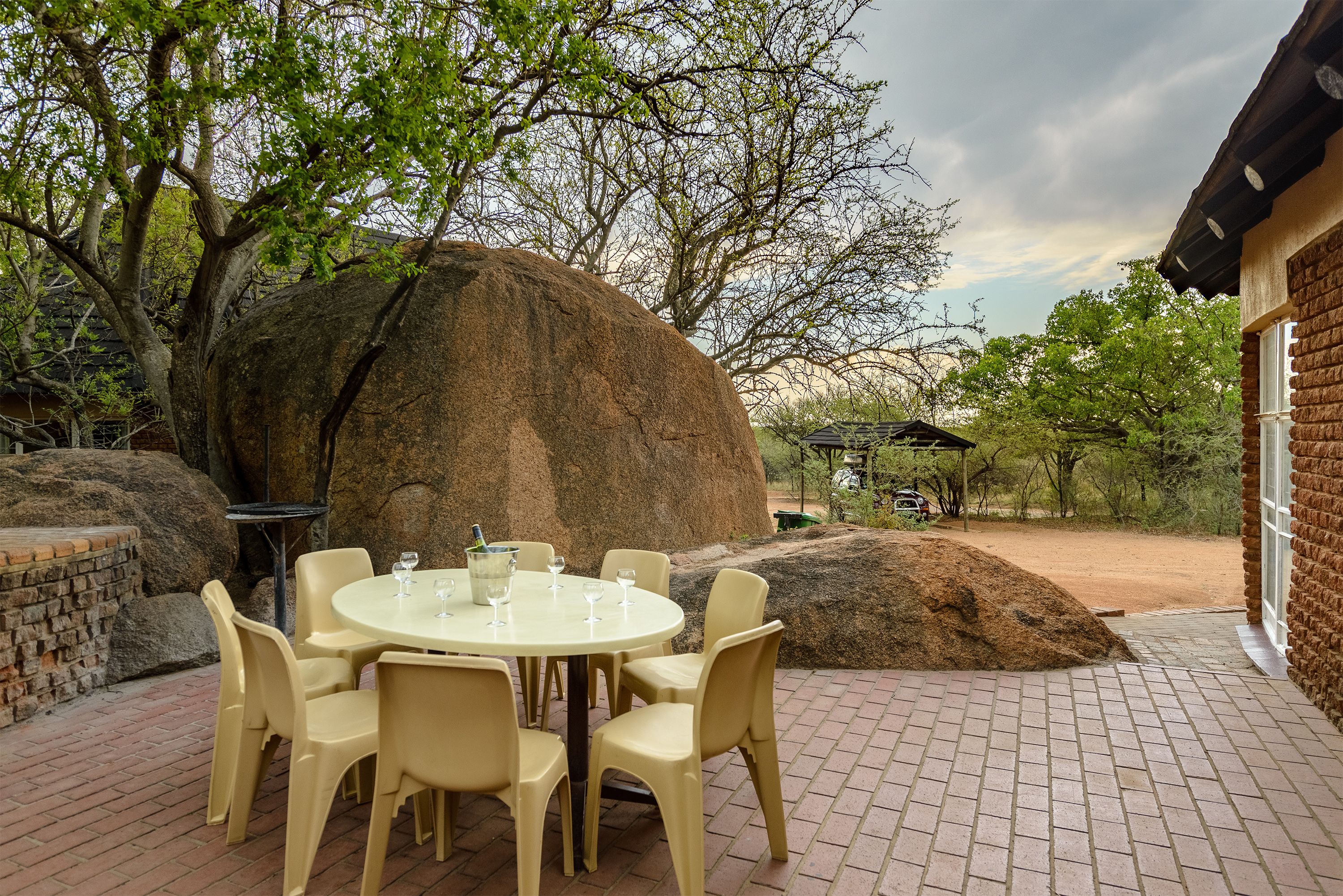 A round table with chairs and a bottle of champagne, set outdoors with a large rock and trees in the background.