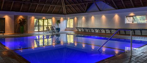 An indoor swimming pool with a blue-lit edge, a wooden ceiling, and a seating area with a view of the outdoors.