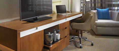 A modern hotel room with a wooden desk, a flat-screen TV, a laptop, a coffee maker, and a comfortable armchair.