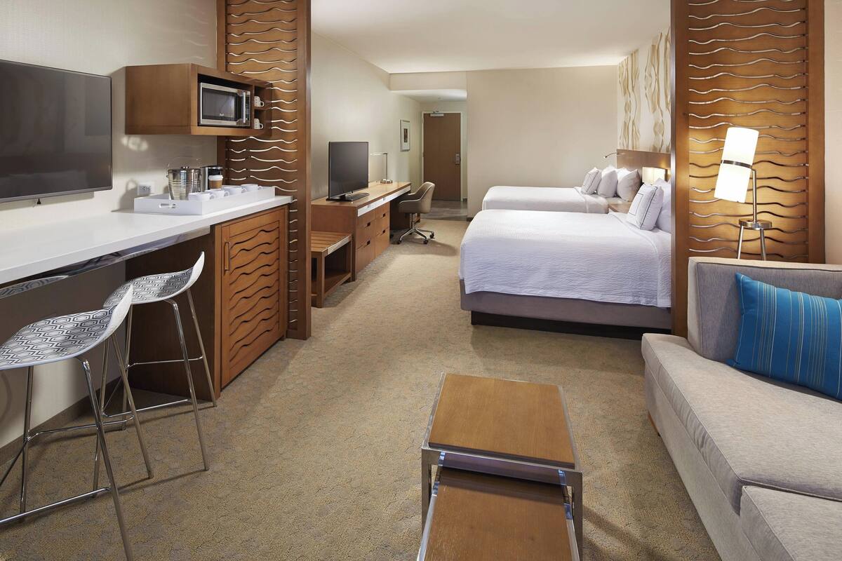 Quarto de hotel com cama, escrivaninha com computador, televisão e área de estar com cadeiras.