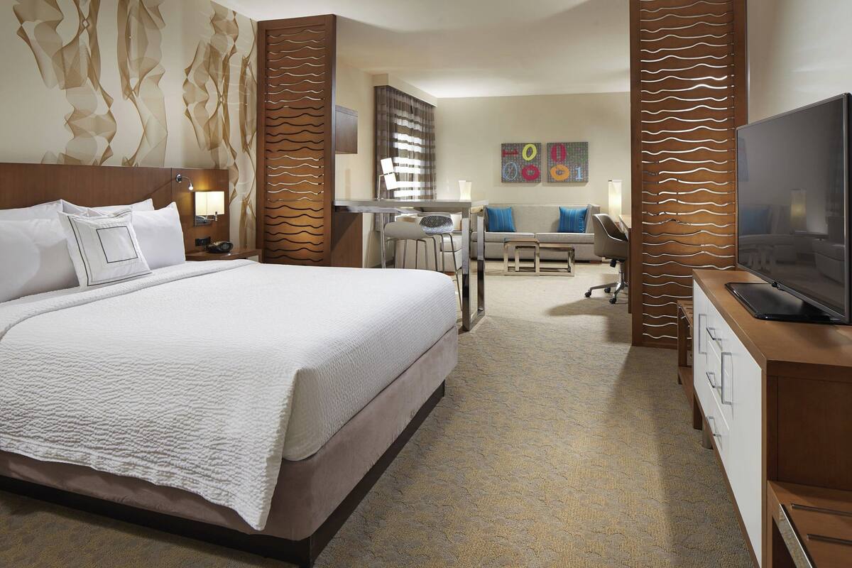 Quarto de hotel com uma cama grande, uma escrivaninha com cadeira, televisão e um sofá.