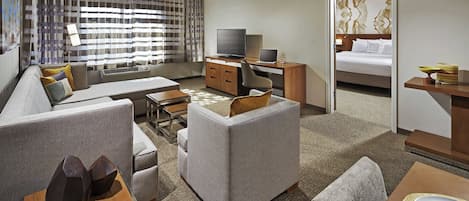 A modern hotel room with a sofa, a dining table, a TV, a laptop, and a bed.