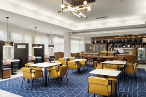 Un moderno vestíbulo de hotel con sillas y mesas amarillas, y una alfombra azul estampada. Se observan luces colgantes y una zona de bar con un panel de menú.