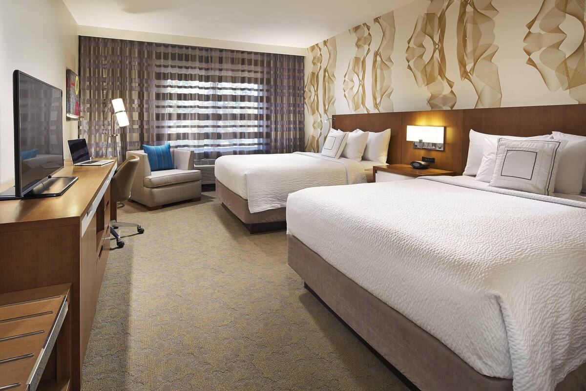 Quarto de hotel com duas camas, uma secretária com um computador portátil, uma televisão e uma janela grande com cortinas.