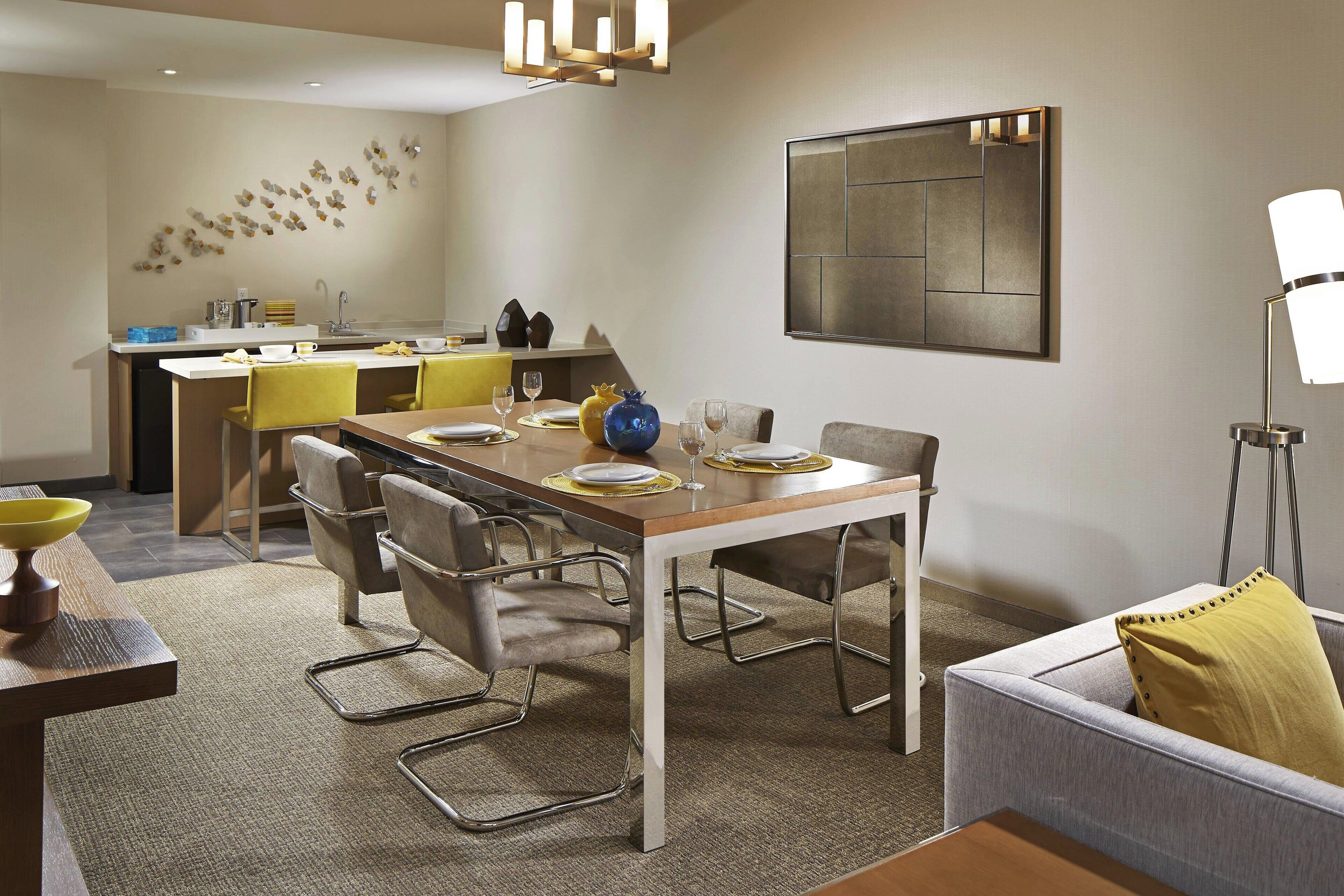 A modern dining area with a wooden table, metal chairs, and a grey sofa. A kitchen is visible in the background with a yellow countertop.