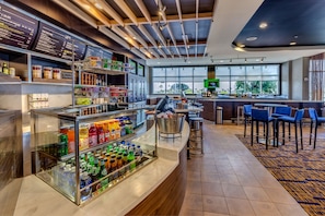 A modern bar area with a glass display case of beverages, a counter with a coffee machine, and a seating area with blue chairs. - Courtyard St. Petersburg Clearwater/Madeira Beach (Madeira Beach)