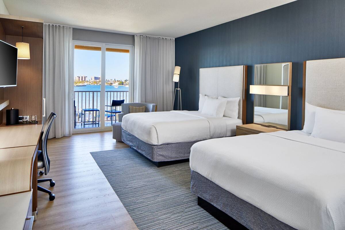 A hotel room with two beds, a desk, a chair, a TV, and a balcony with a view.