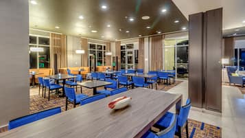 Un espace de restauration moderne dans un hôtel, avec de nombreuses tables et chaises, une grande table en bois agrémentée d’un objet décoratif, et une vue sur la ville la nuit, grâce à de grandes fenêtres.