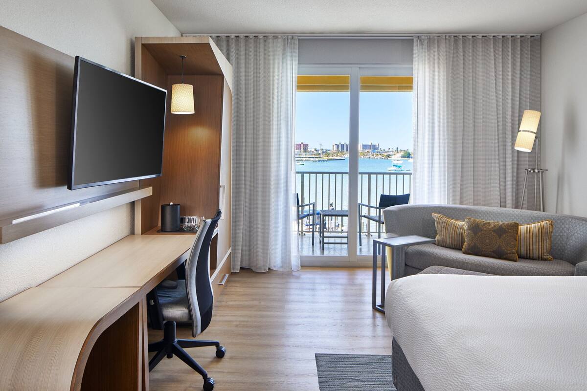 A hotel room with a bed, a desk, a television, a balcony with a view of the city and water.