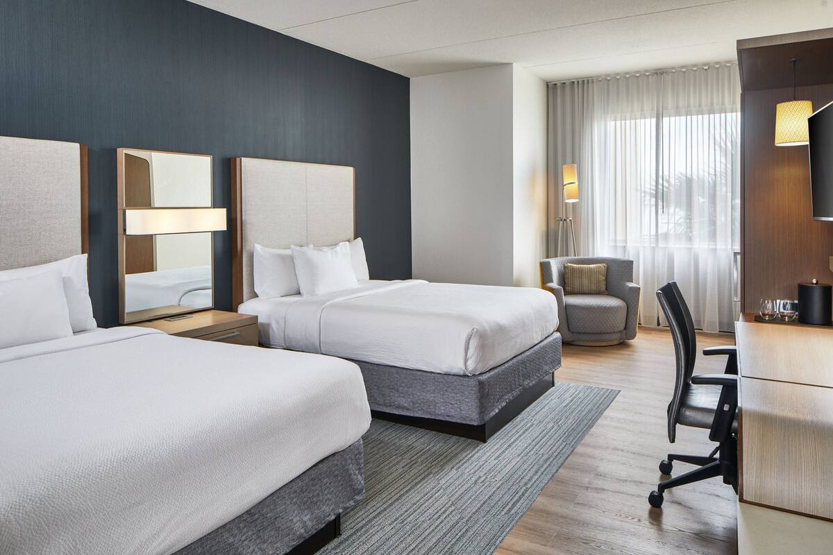 A hotel room with two beds, a desk, a chair, a lamp, and a large window with curtains.