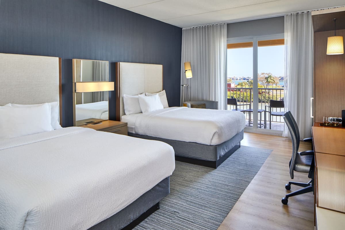 A hotel room with two beds, a desk, and a chair. There is a large window with a view of the outdoors.
