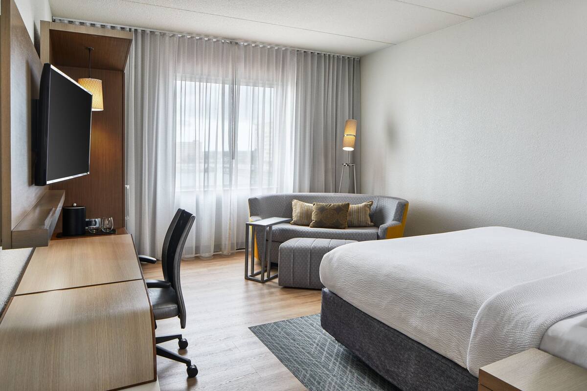A modern hotel room with a large bed, a desk, a chair, a sofa, a TV, and a window with curtains.