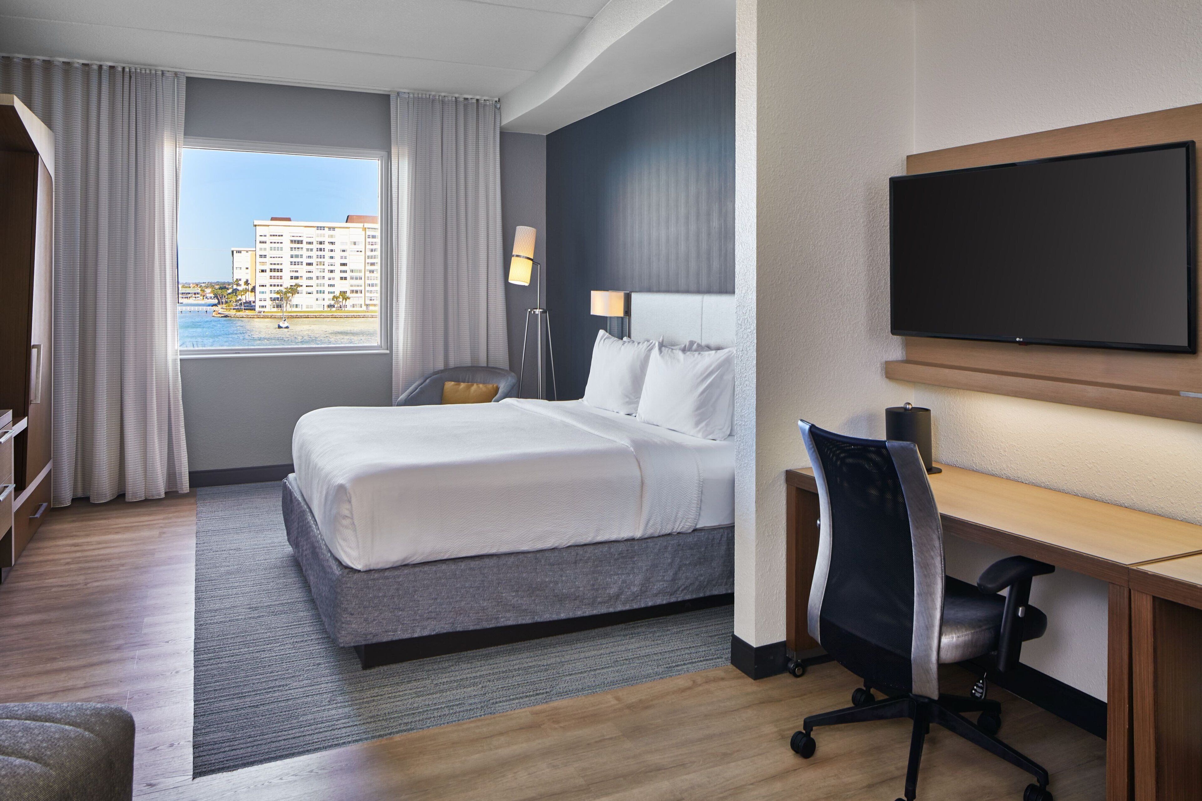 A hotel room with a bed, desk, chair, TV, and a view of a building through the window.