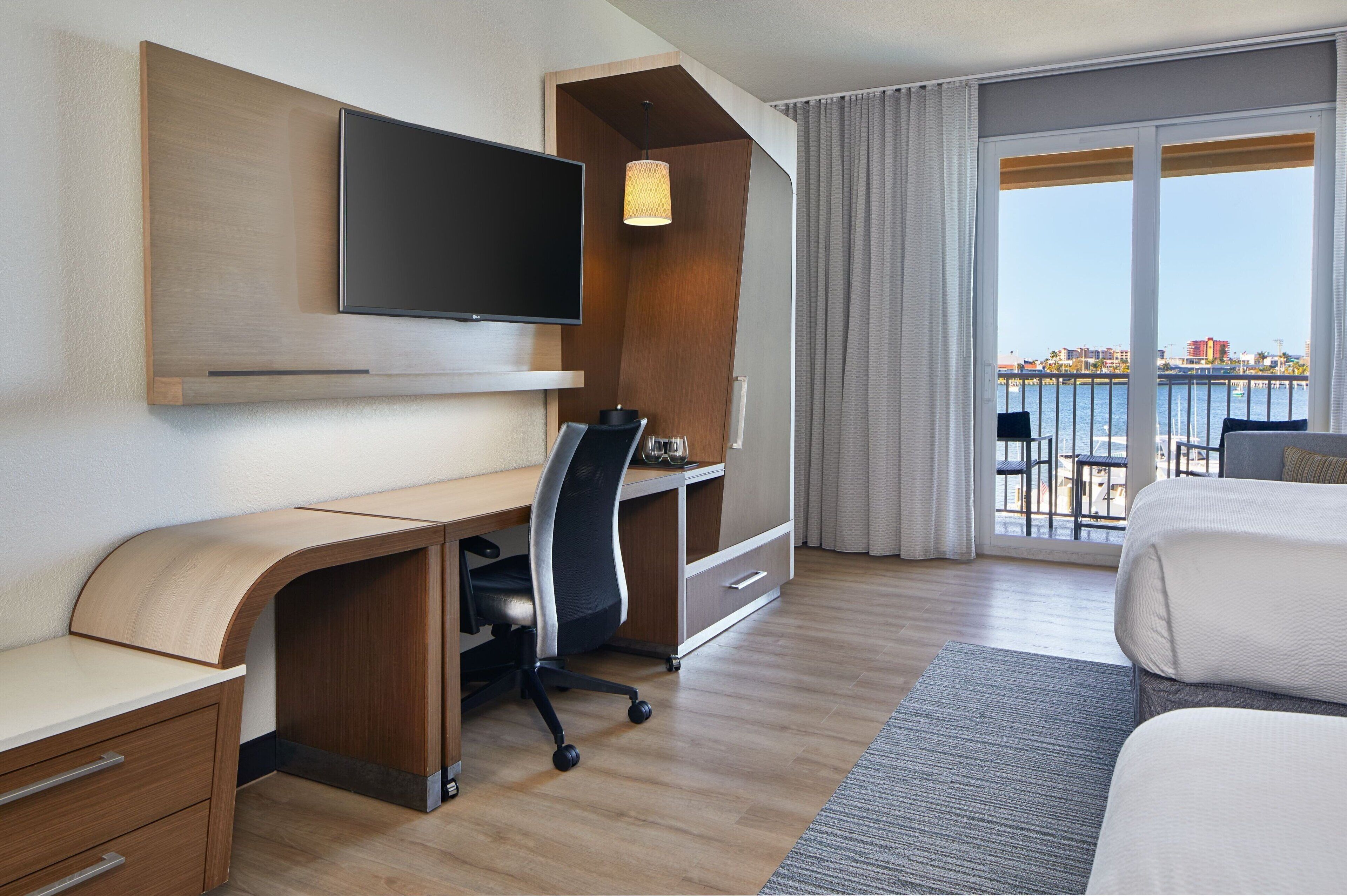A hotel room with a wooden desk, a flat-screen TV, a bed, and a balcony with a city view.