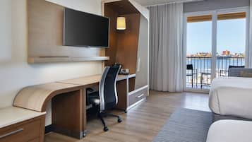A hotel room with a wooden desk, a flat-screen TV, a bed, and a balcony with a city view.