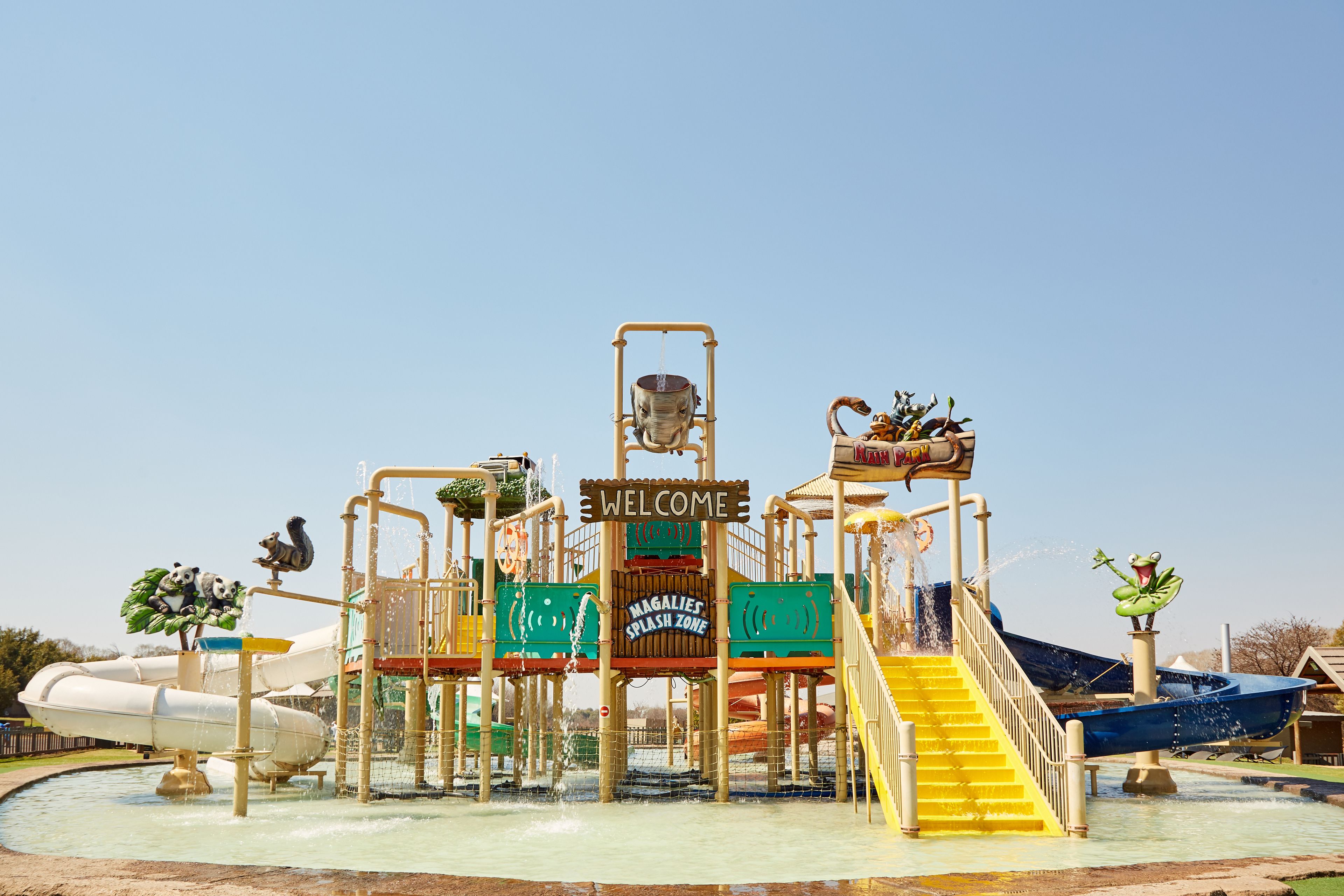Un parco acquatico con un cartello che dice 'Benvenuti', uno scivolo e un’area giochi con getti d’acqua.