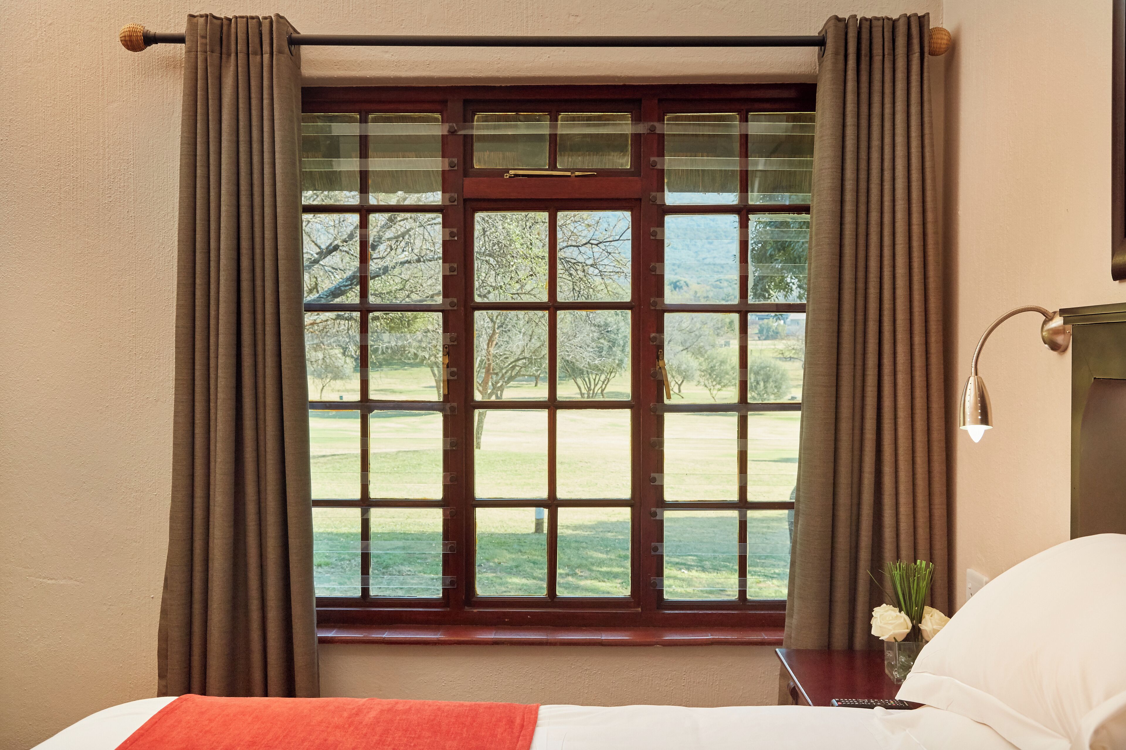 Habitación de hotel con un ventanal grande, cortinas, una mesita de noche con un florero y una cama con una colcha roja.