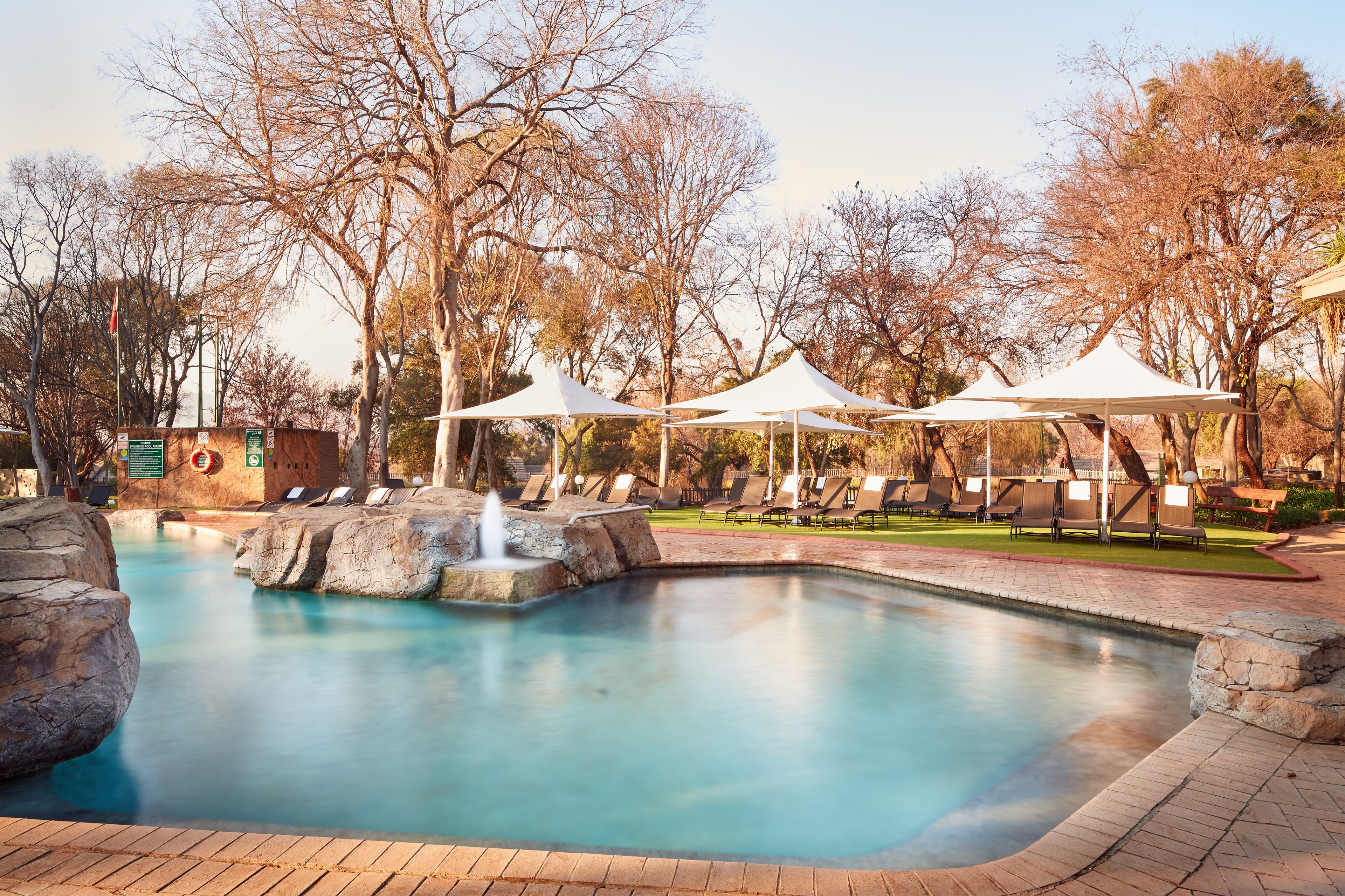 Une piscine agrémentée d’une fontaine, entourée de chaises longues et de parasols blancs.