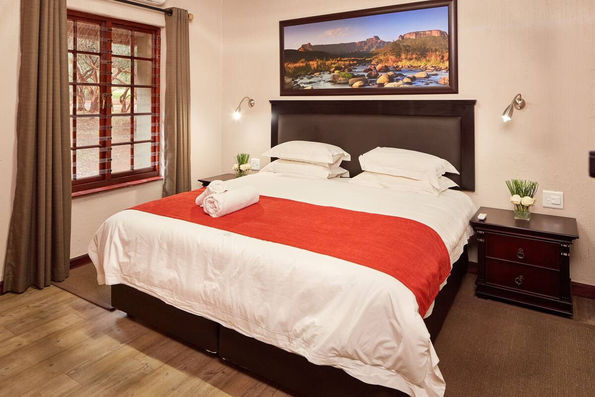 Une chambre d’hôtel avec un grand lit, une table de chevet ornée d’un vase de fleurs, un tableau au-dessus du lit et une fenêtre avec des rideaux.